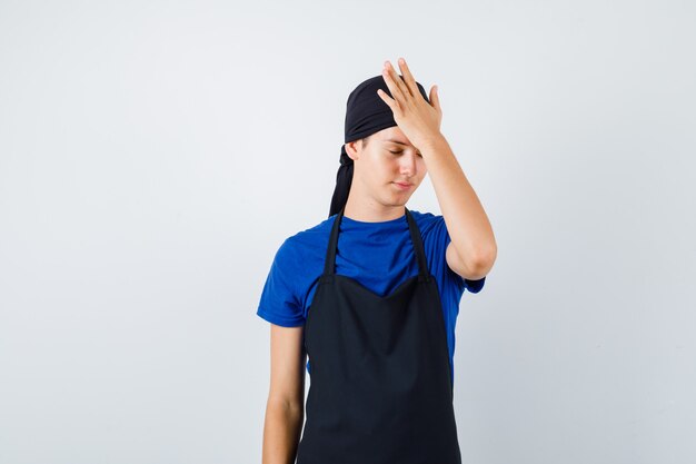 Portrait of young teen cook with hand on head in t-shirt, apron and looking forgetful front view