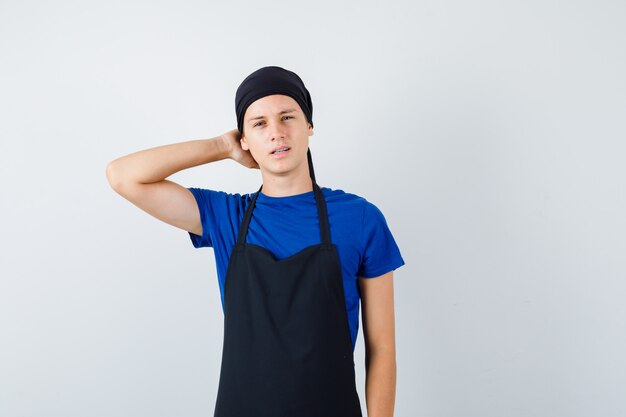 Portrait of young teen cook with hand behind head in t-shirt, apron and looking pensive front view