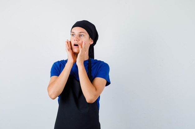 Portrait of young teen cook with hands near open mouth in t-shirt, apron and looking shocked front view