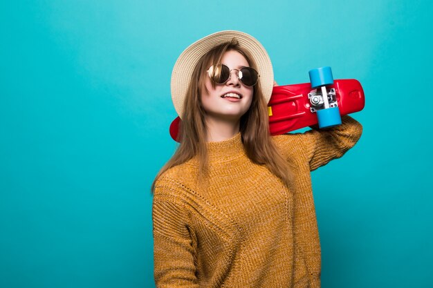 Portrait of young teen woman in sunglasses and hat holding skateboard while standing isolated over green wall