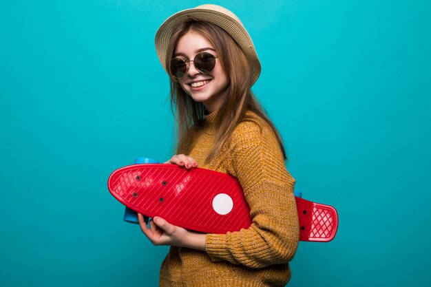 Portrait of young teen woman in sunglasses and hat holding skateboard while standing isolated over green wall