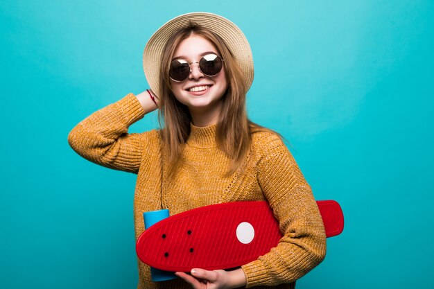 Portrait of young teen woman in sunglasses and hat holding skateboard while standing isolated over green wall