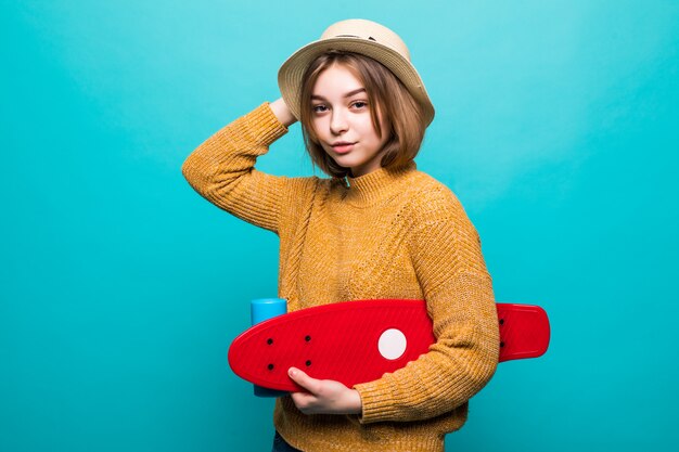 Portrait of young teen woman in sunglasses and hat holding skateboard while standing isolated over green wall