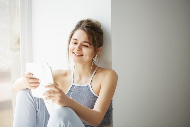 白い壁の上の窓の近くに座ってインターネットを閲覧するタブレットサーフィンwebを見て笑ってヘッドフォンで若い10代の陽気な女性の肖像画。