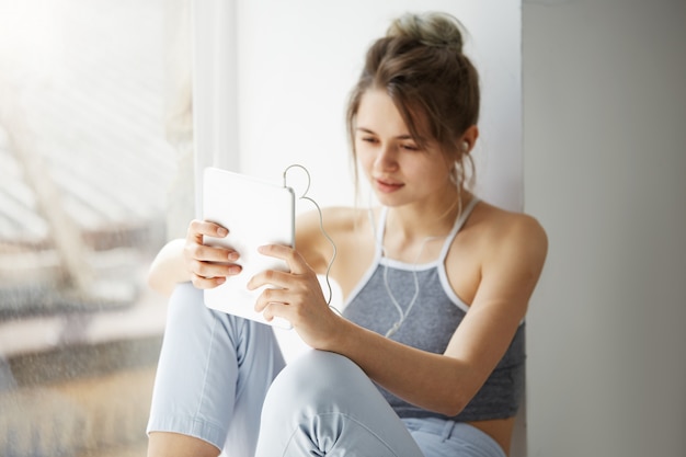 Portrait of young teenage cheerful woman in headphones smiling looking at tablet surfing web browsing internet sitting near window over white wall.