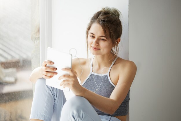 Portrait of young teenage cheerful woman in headphones smiling looking at tablet surfing web browsing internet sitting near window over white wall.