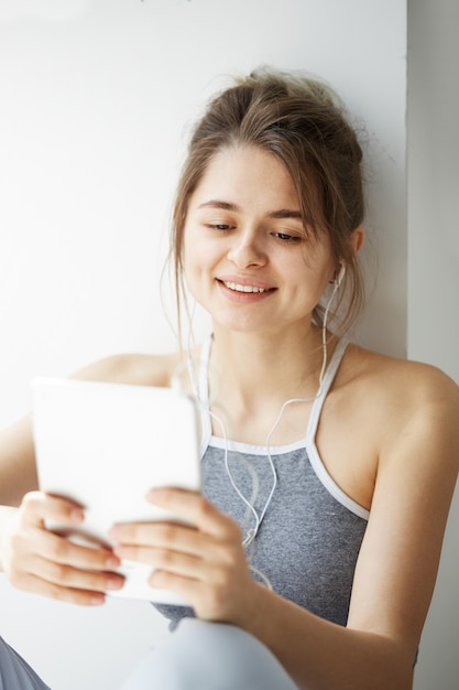 Portrait of young teenage cheerful woman in headphones smiling looking at tablet surfing web browsing internet sitting near window over white wall.