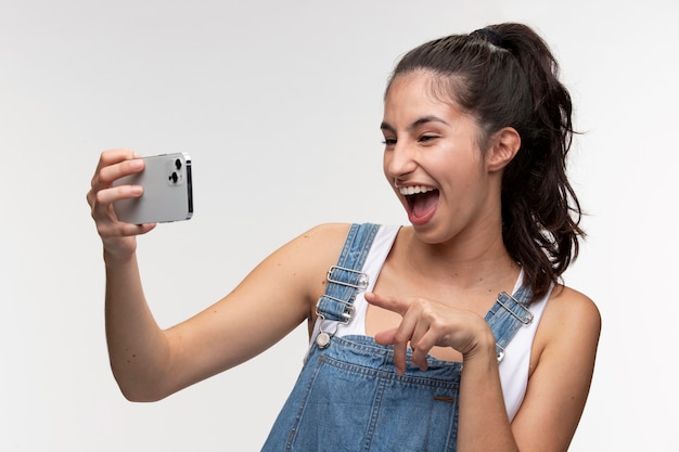 Free photo portrait of young teenage girl in overalls taking a selfie with smartphone