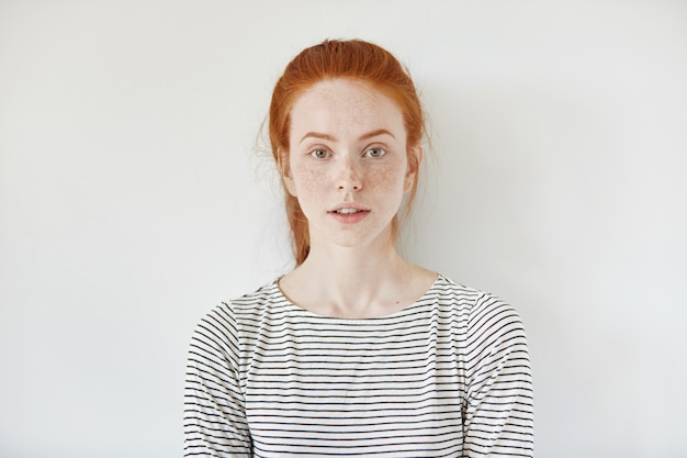 Portrait of young tender redhead teenage girl with healthy freckled skin wearing striped top looking with serious or pensive expression. Caucasian woman model with ginger hair posing indoors