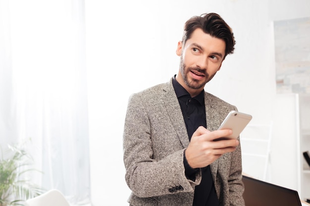 Портрет молодого задумчивого бизнесмена в черной рубашке и куртке, стоящего и мечтательно смотрящего в сторону, держа в руке мобильный телефон в изолированном офисе