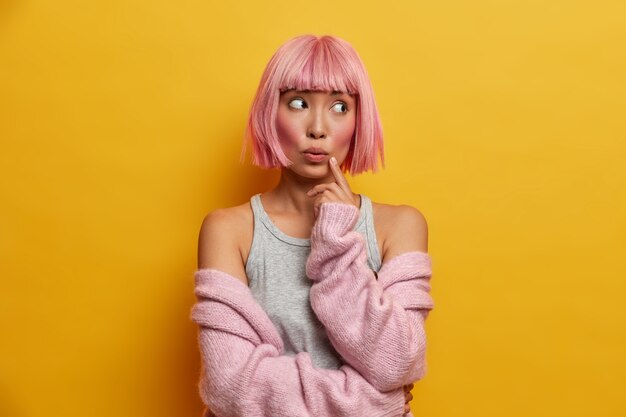 Portrait of young thoughtful surprised asian woman with rosy hair, keeps finger on face and looks wondered aside, wears warm knitted sweater