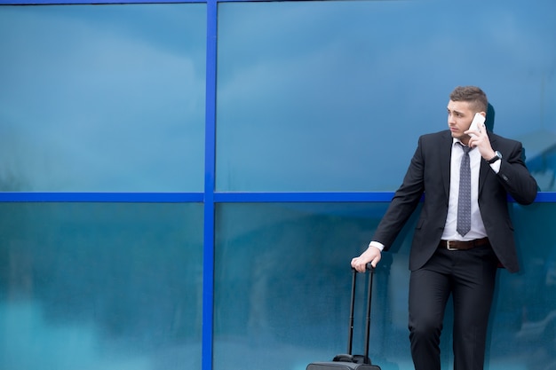 Portrait of young traveler in suit standing with suitcase and making call. Copy space