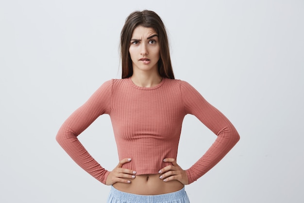 Portrait of young unhappy caucasian girl with dark long hair in trendy pink top and sporty blue trousers looking  with angry expression, holding hands on waist, seeing her boyfriend