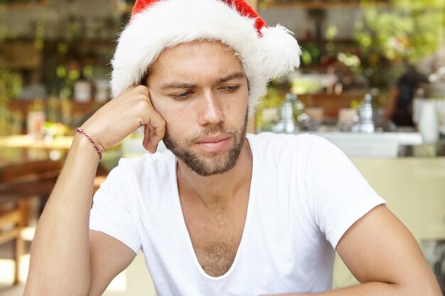 Portrait of young unshaven man looking down with unhappy face, sitting at cafe alone, leaning on his elbow