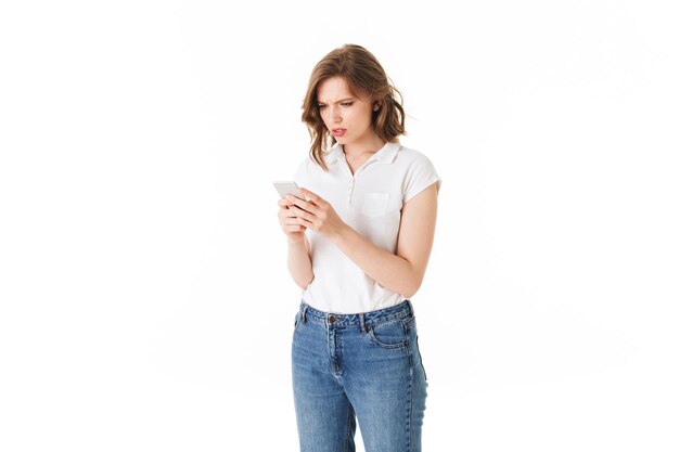 고립 된 흰색 배경에 손에 핸드폰을 들고 서 있는 티셔츠와 청바지에 젊은 화가 여자의 초상화