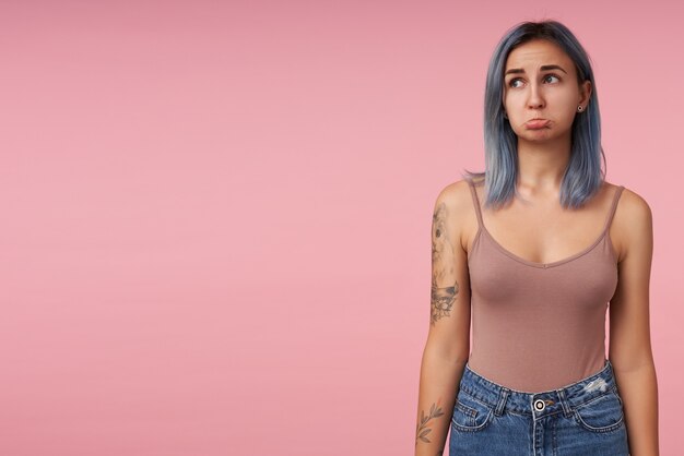 Free photo portrait of young upset short haired tattooed lady twisting sadly her mouth while looking aside and keeping hands along body, standing on pink