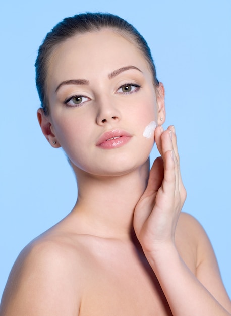 Portrait of young woman applying cream on her beautiful fresh face on blue