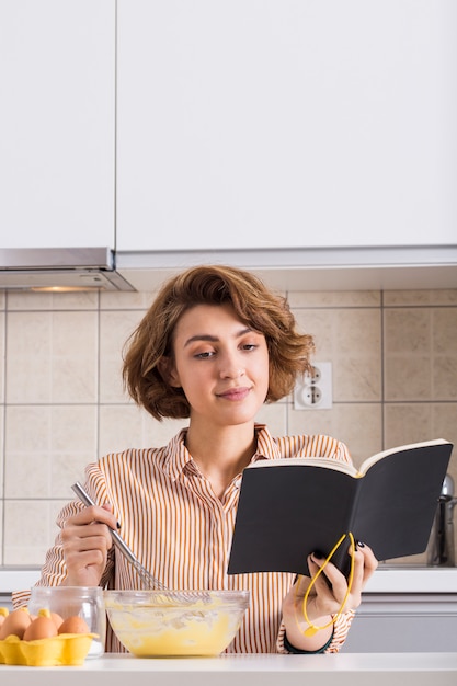 Free photo portrait of a young woman beating the eggs while reading the recipe book