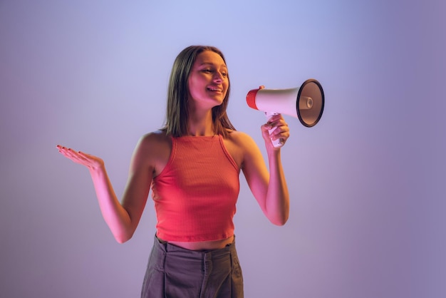 Portrait of young woman in casual cloth shouting in megaphone isolated over purple studio background in neon ligh