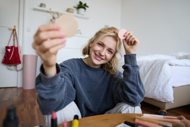 Free photo portrait of young woman content creator showing beauty makeup products at camera sitting on floor in