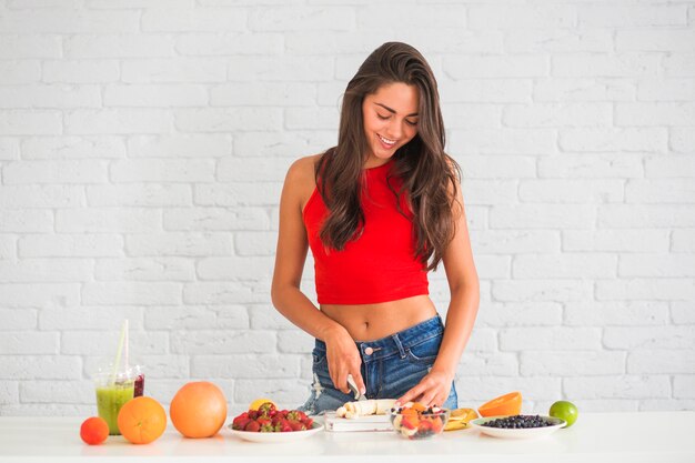 Portrait of young woman cutting banana standing against white brick wall