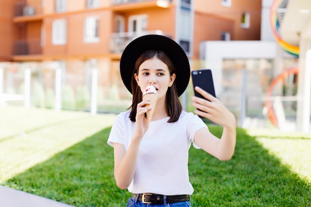 アイスクリームを食べると夏の通りでカメラでselfie写真を撮る若い女性の肖像画。