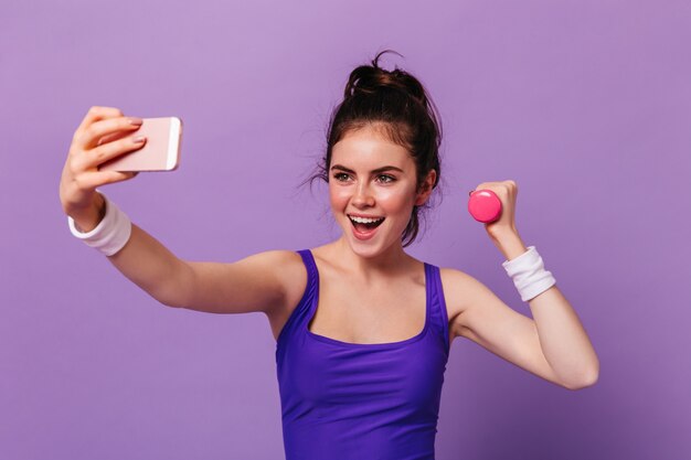 Portrait of young woman in fitness top holding pink dumbbell and taking selfie on purple wall
