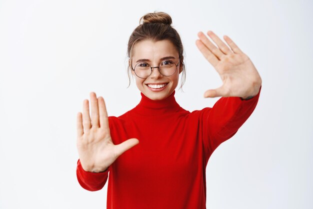 Portrait of young woman in glasses stretch out hand frames and smiling picturing a moment imaging something standing against white background