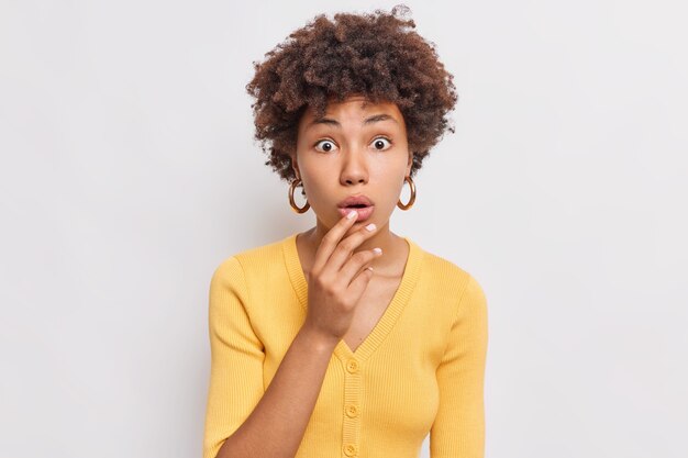 Portrait of young woman has startled expression raises eyebrows holds breath dressed in yellow jumper cannot believe her eyes isolated over white wall