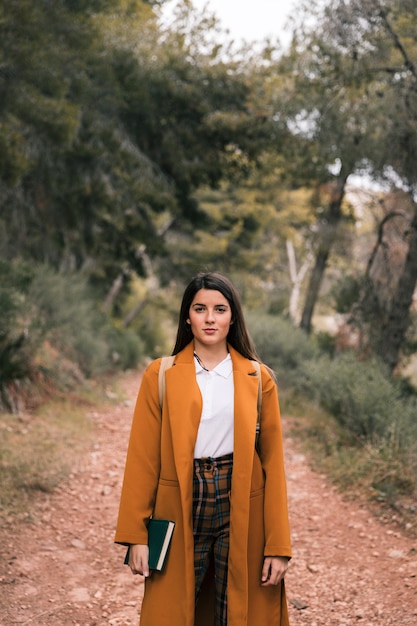 Free photo portrait of a young woman holding book in hand standing on forest trail