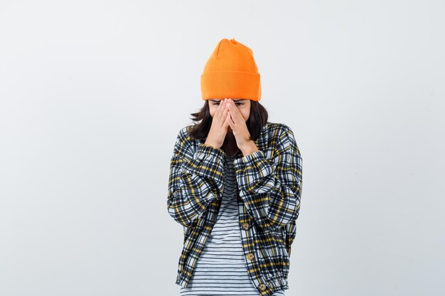 Portrait of young woman holding face with palms in orange hat and checkered shirt