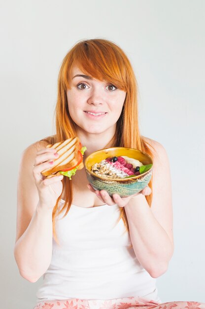 Portrait of young woman holding sandwich and oatmeal bowl against white background