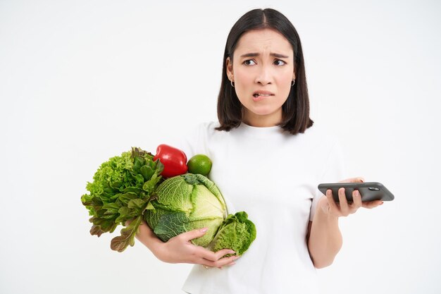 Portrait of young woman holds vegetables organic food holds smartphone with concerned worried face i