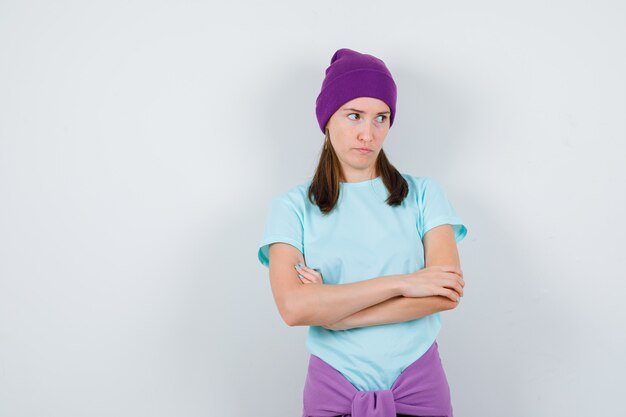 Portrait of young woman keeping arms folded, looking away in t-shirt, beanie and looking dismal front view