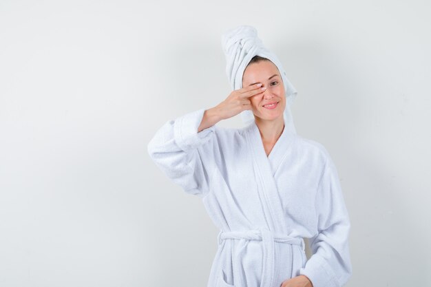 Portrait of young woman keeping fingers on eye in white bathrobe, towel and looking cheery front view