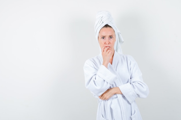 Portrait of young woman keeping hand on chin in white bathrobe, towel and looking anxious front view