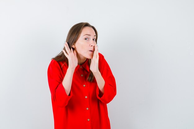 Портрет молодой женщины, держащей руку у рта, подслушивая секрет в красной блузке и глядя на любопытный вид спереди