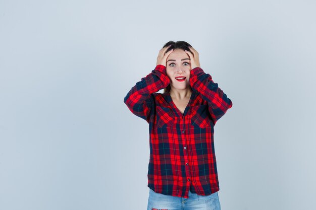 Portrait of young woman keeping hands on head in checked shirt and looking excited front view