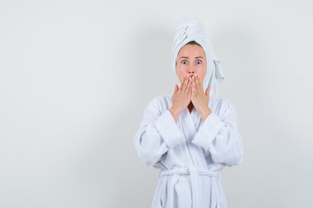 Portrait of young woman keeping hands on mouth in white bathrobe, towel and looking surprised front view