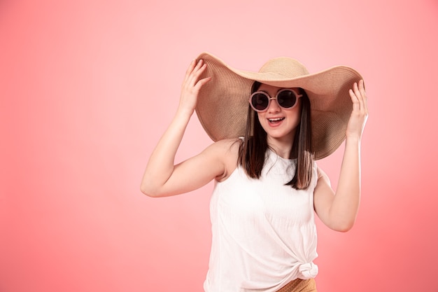 大きな夏の帽子とメガネ、ピンクの背景の若い女性の肖像画。夏のコンセプトです。