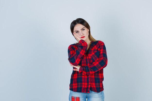 Portrait of young woman leaning cheek on palm in checked shirt and looking dismal front view