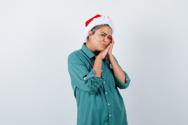 Portrait of young woman leaning on palms as pillow in shirt, Santa hat and looking peaceful front view