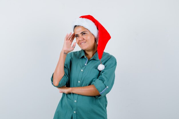 Portrait of young woman looking away while thinking, biting lip in shirt, Santa hat and looking positive front view