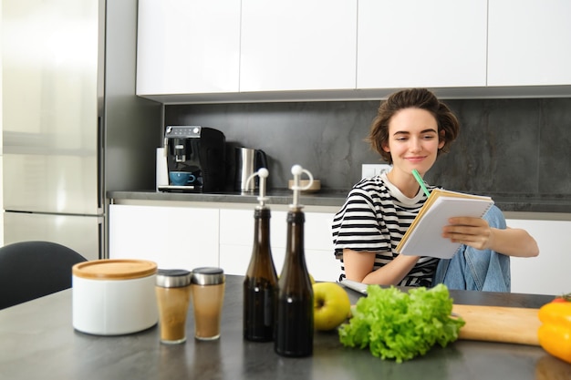 Free photo portrait of young woman looking at cooking ingredients on kitchen counter and making notes writing