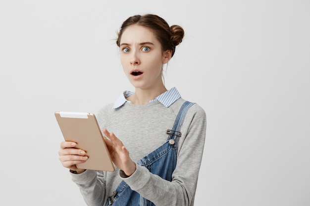 Portrait of young woman looking  expressing tension and fear holding golden tablet. Frustrated female receptionist realizing she confused her boss schedule. Negative emotions