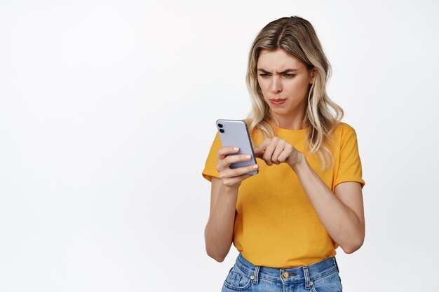 Portrait of young woman looking serious at mobile phone, poking smartphone screen and frowning concentrated, standing on white