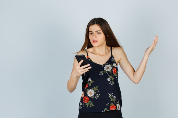 Portrait of young woman looking at smartphone in blouse, skirt and looking nervous front view