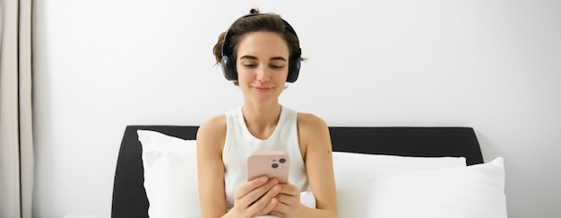 Free photo portrait of young woman lying in bed wearing wireless headphones looking at smartphone watching