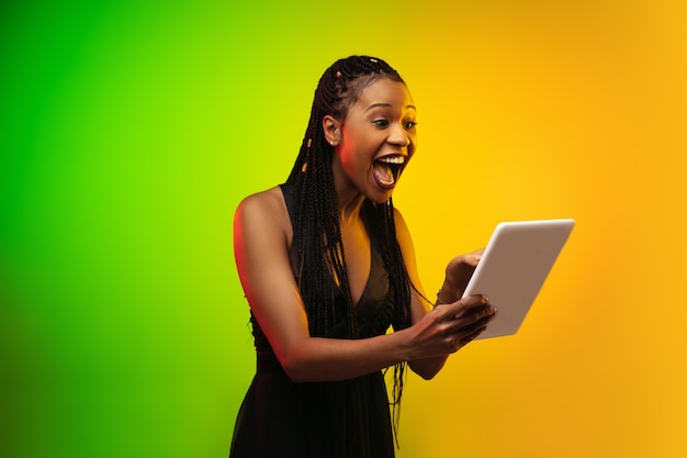 Portrait of young woman in neon light on gradient backgound. Holding a tablet.