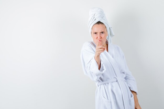 Portrait of young woman pointing at camera in white bathrobe, towel and looking angry front view